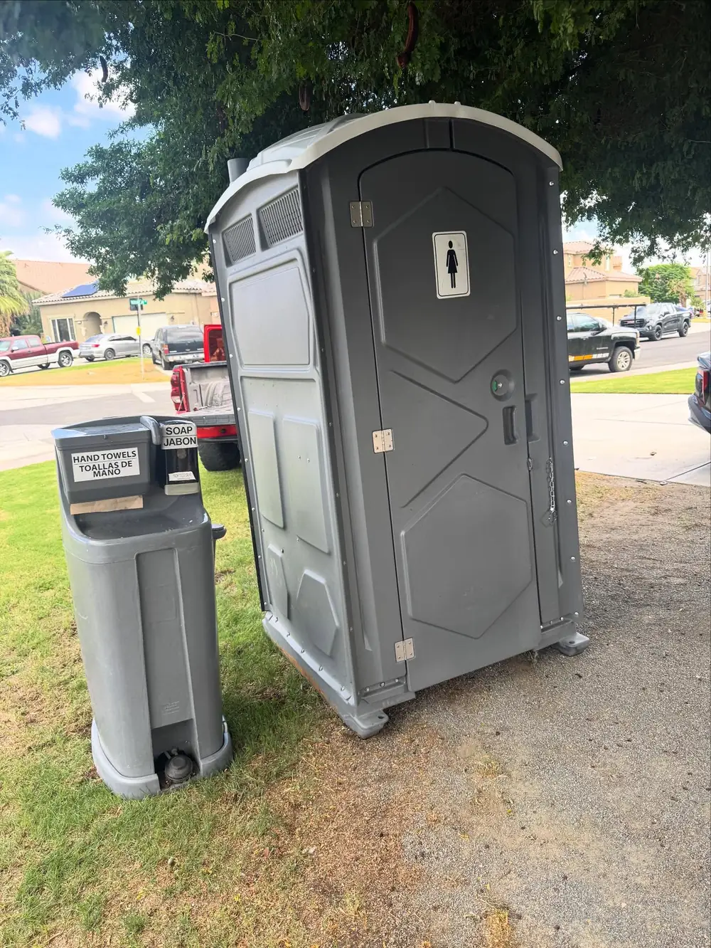 Grey porta potty next to a portable hand-washing station on a lawn