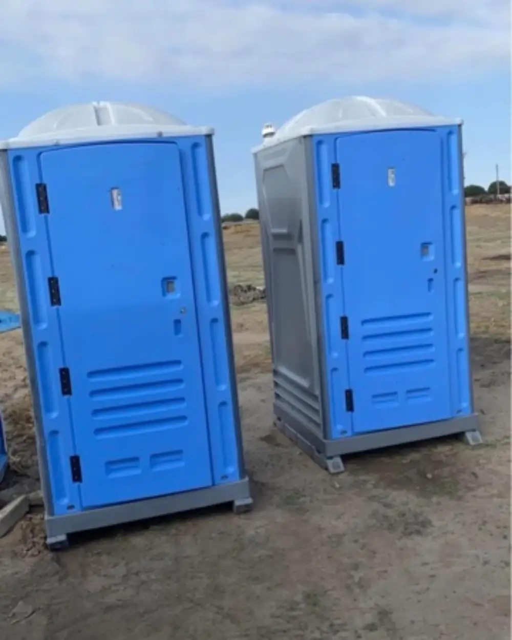 Two blue standard porta potties set side by side on a dirt jobsite